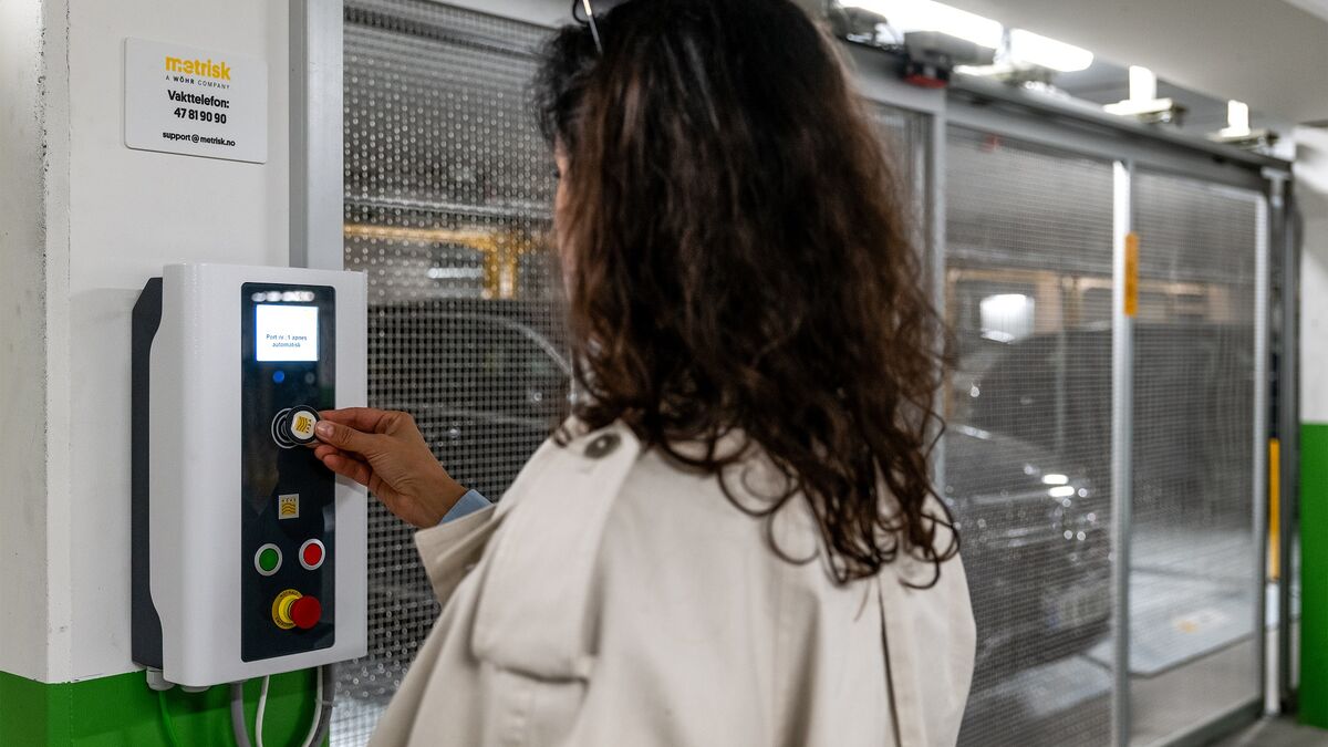 A person operates the control panel of a semi-automatic parking system to retrieve a parked vehicle. In the background, the movable parking spaces and a vehicle within the parking facility are visible.
