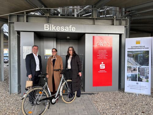 Three people are standing with a bicycle in front of the entrance to the automated bicycle parking system